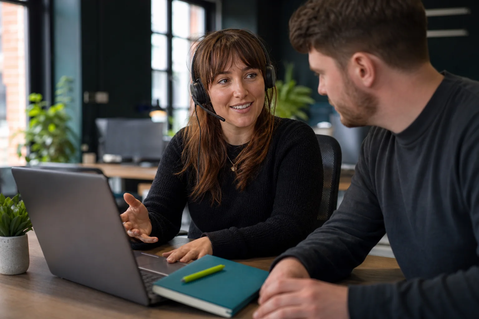 A headset-wearing colleague discussing a laptop with a coworker